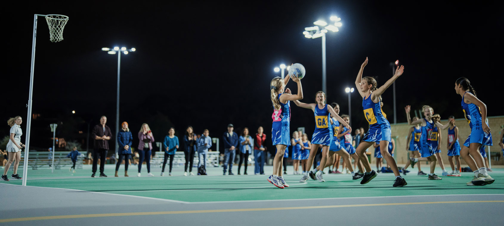 A netball game in progress at night being lit by floodlights with parents watching on the sidelines